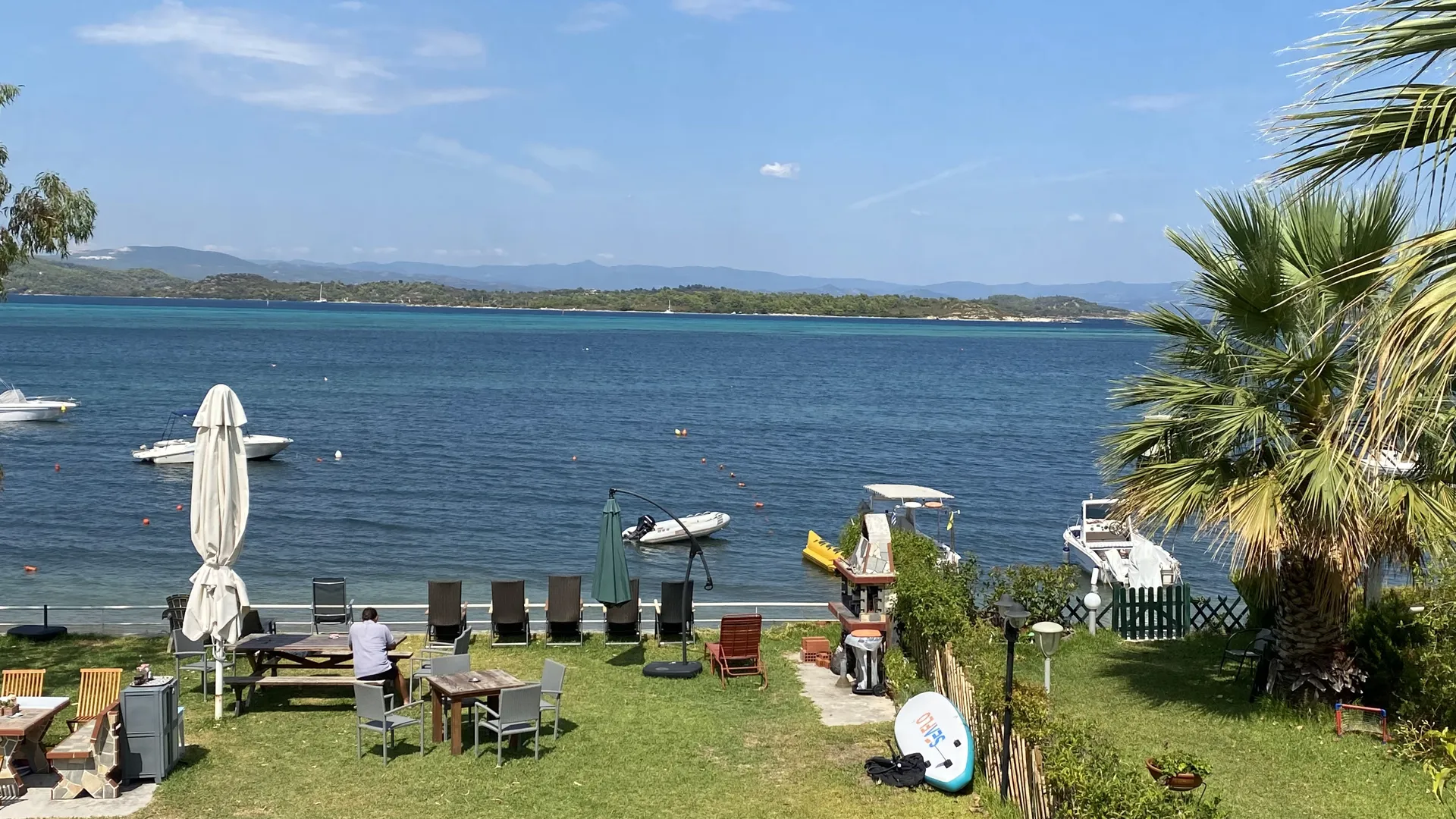 The seaside garden of Studios EDRA with umbrellas and deck chairs on the lawn, boats moored on the turquoise bay of Vourvourou